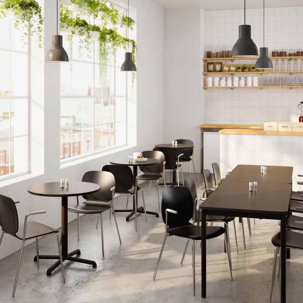 Une salle à manger avec des chaises et des tables de tailles différentes, des lampes HEKTAR noires suspendues au-dessus.