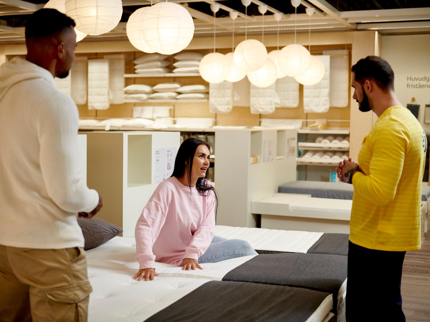 Osoba sedící na posteli v obchodním domě IKEA, vybírající si novou matraci, se kterou ji radí zaměstnanec IKEA.