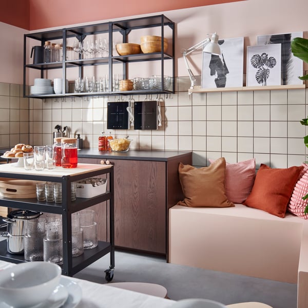 A brown kitchen in a left back corner with open shelves on the wall and beige tiles under them. A black kitchen island with a wooden top is standing in front of the kitchen. To the left is a pink bench with cushions in different colours.