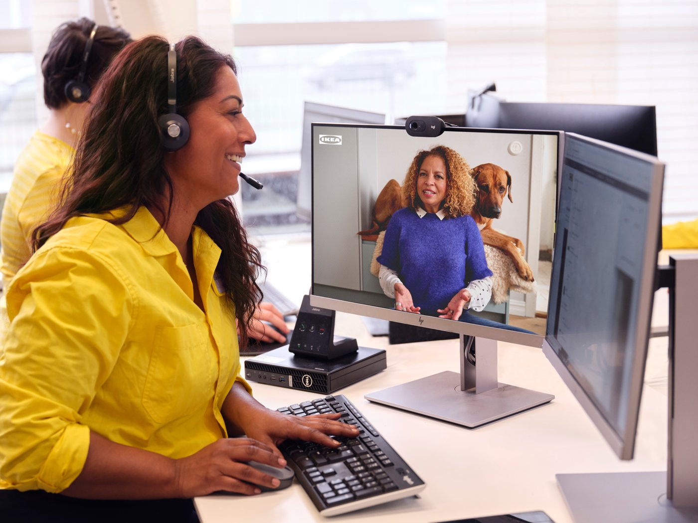An IKEA co-worker has a headset on with a yellow shirt on and is speaking to a customer virtually on the computer.