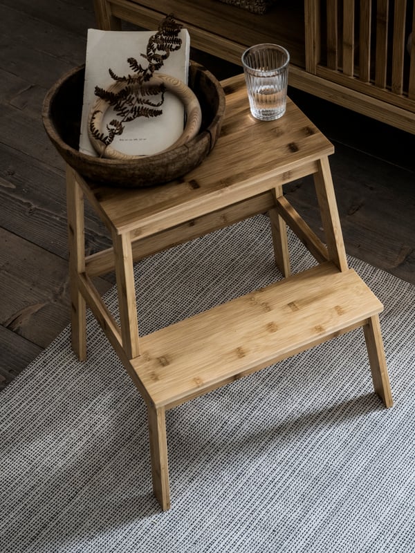 سلم متدرج TENHULT عليه كوب من الماء ووعاء خشبي يحتوي على كتاب وحلقة خشبية وبعض أوراق الشجر الجافة.