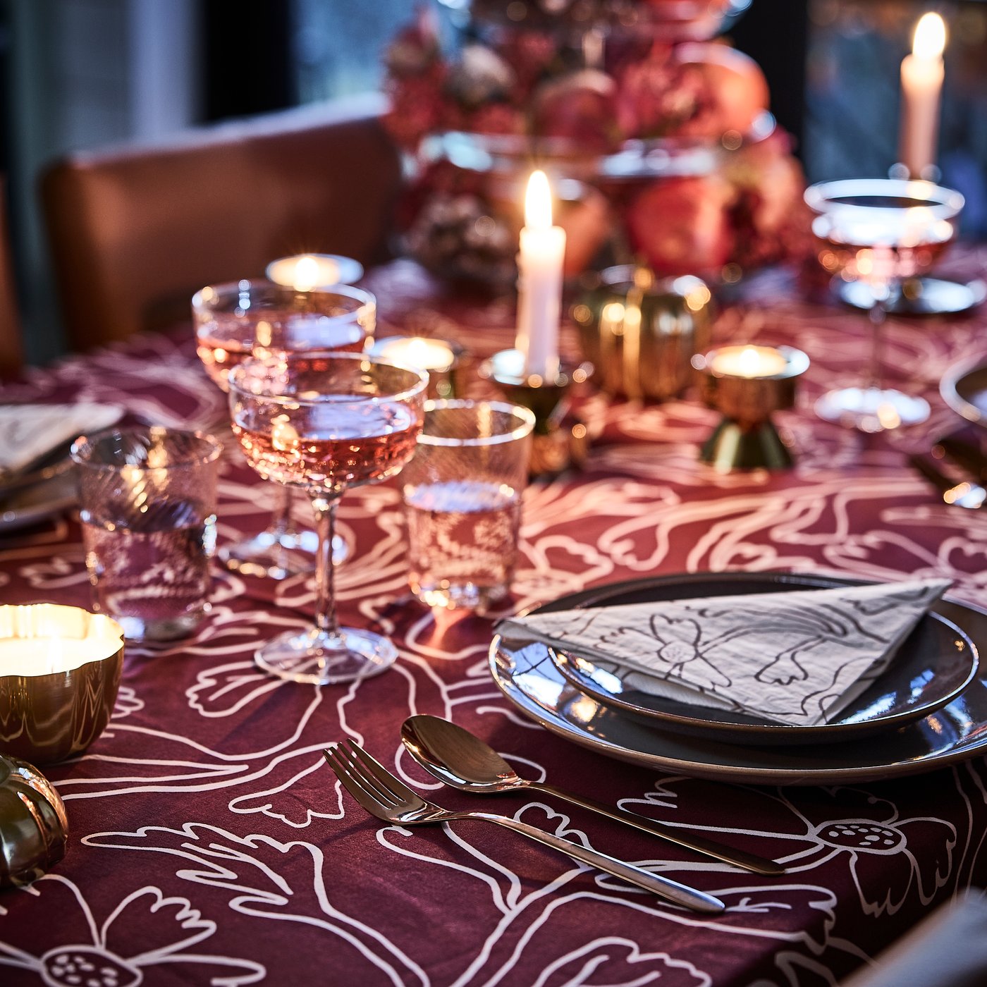 Una tavola nei toni del rosa e del rosso, con candele accese, una tovaglia HÖSTAGILLE rossa fantasia, tovaglioli di carta e coppe da champagne.