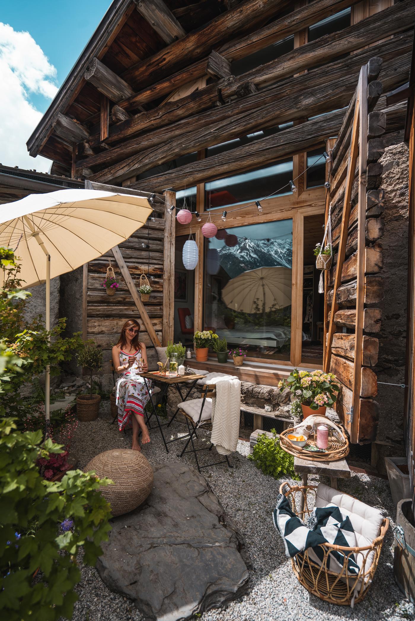 Rustikale Terrasse mit Sonnenschirm, Holztisch und Pflanzen vor einem Chalet mit Blick auf die Alpen.