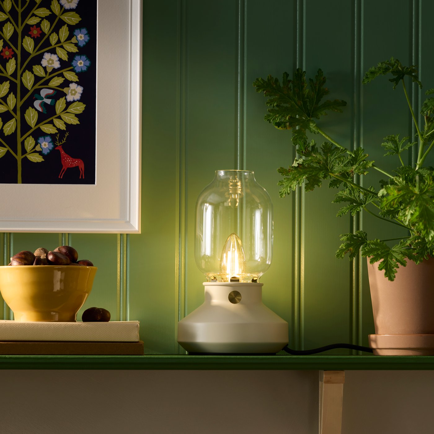 En beige TÄRNABY bordslampa med glaskupa står på en grön hylla mellan en skål med kastanjer och en grön växt.