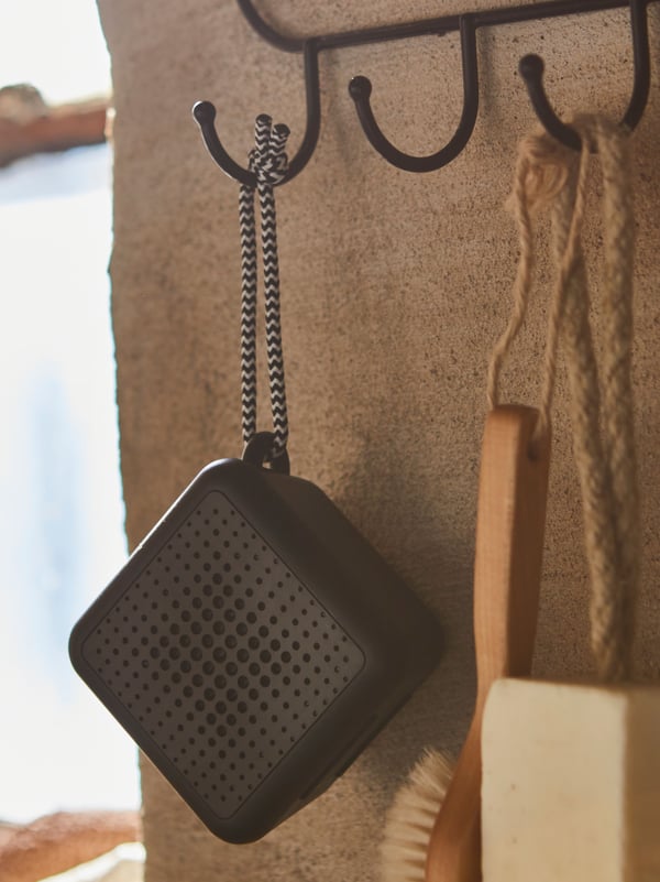 A soap, a brush and a black VAPPEBY portable, waterproof Bluetooth speaker hanging side by side against a brown stone wall.