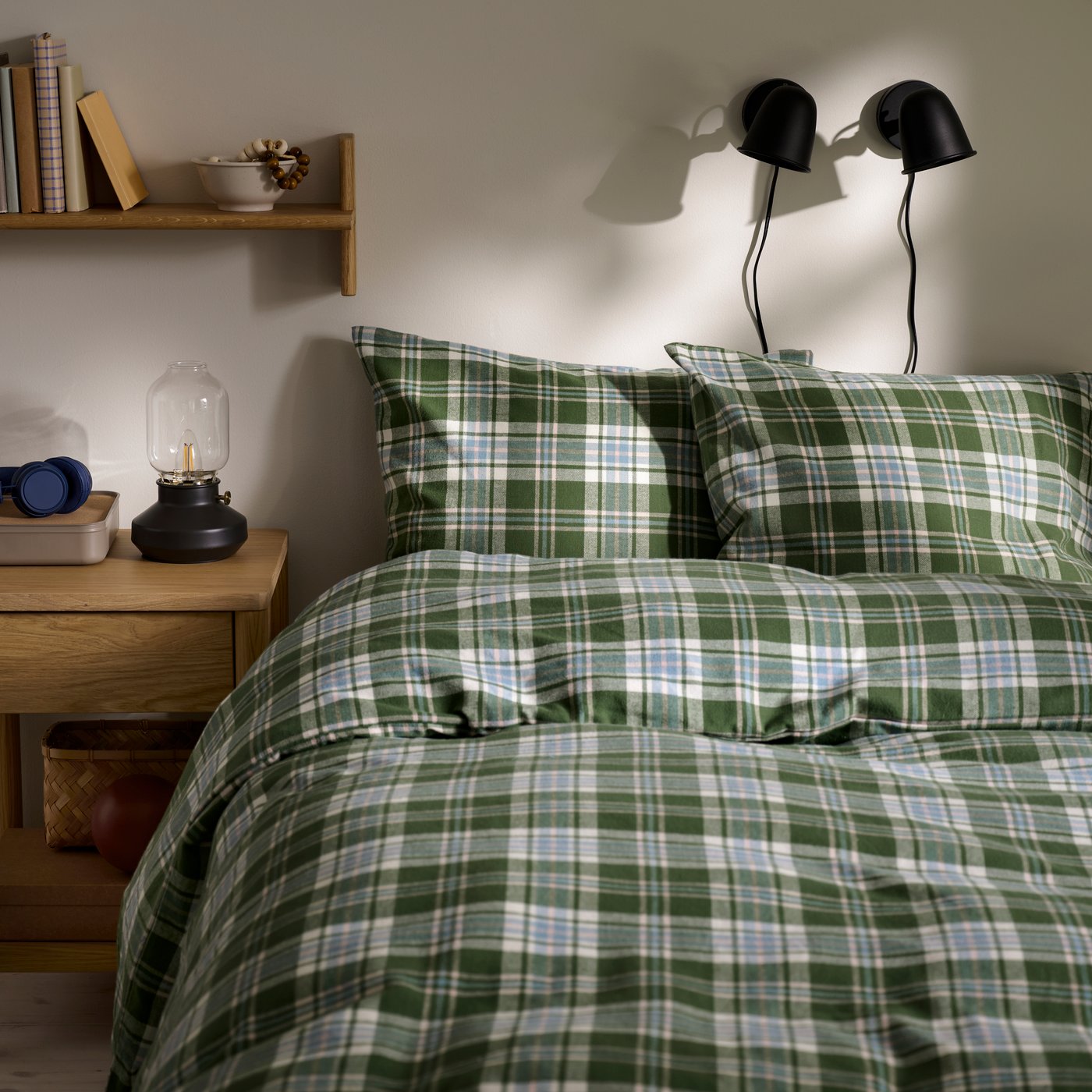 A bedroom with a green checked BANBJÖRNBÄR duvet cover on a bed, along with a wooden bedside table and a black wall lamps.