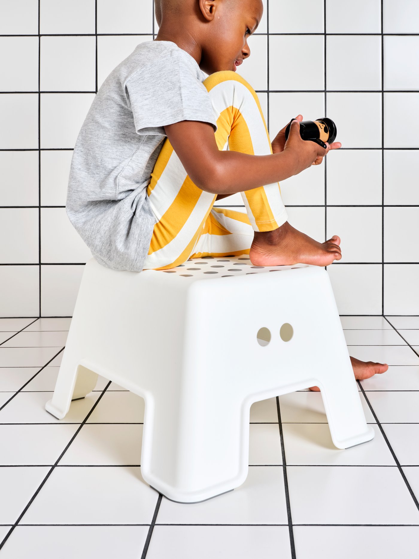 A young child holds a toy while sitting on a white plastic BOLMEN step stool in a white-tiled bathroom.