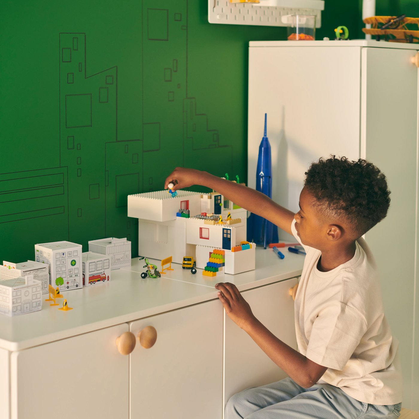 A boy sits on a stool, playing with a BYGGLEK LEGO brick set placed on a white SMÅSTAD cabinet-and-wardrobe combination.