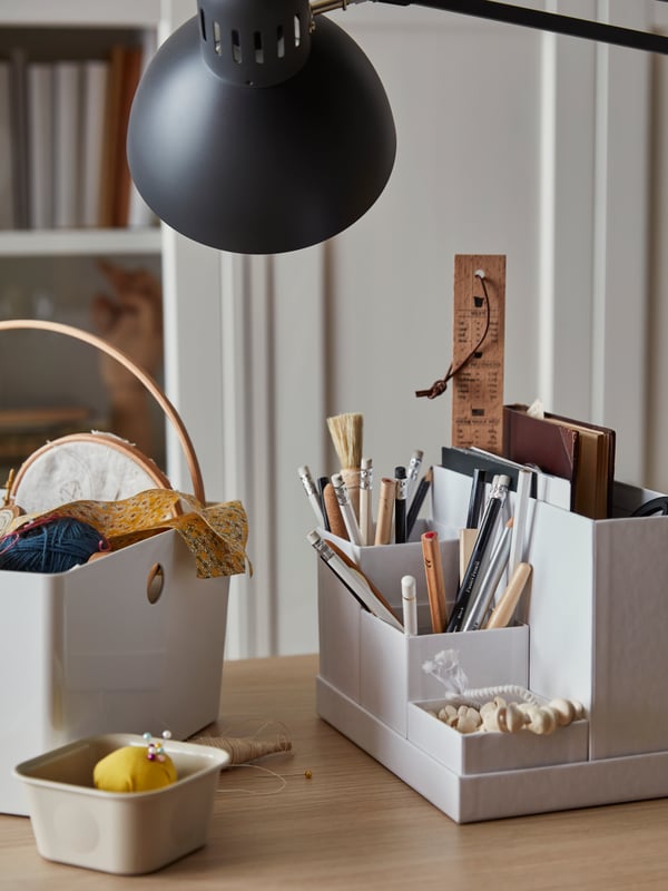 Desktop with sewing material in KUGGIS and NOJIG boxes, writing accessories in a white TJENA organiser, and a TERTIAL lamp.