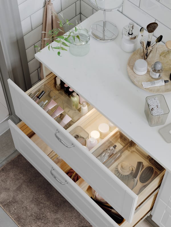 A wash-stand with toiletries on top and two open drawers with GODMORGON boxes with compartments inside containing make-up.