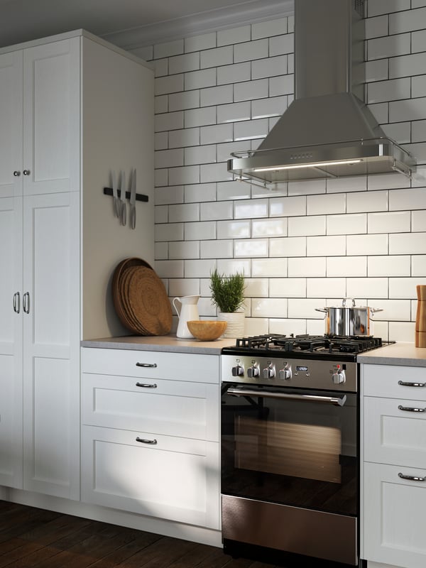 A kitchen with ENKÖPING white fronts, an ALLMÄNBILDAD wall-mounted extractor hood in stainless steel, items on the countertop.