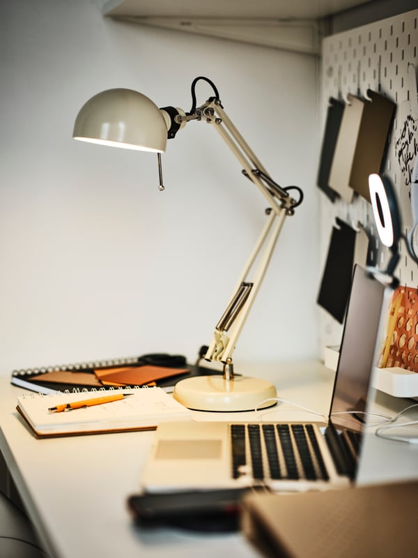 Bež FORSÅ radna lampa, laptop računar, sveska i olovka na beloj ploči stola, plus bela perforirana ploča na zidu.