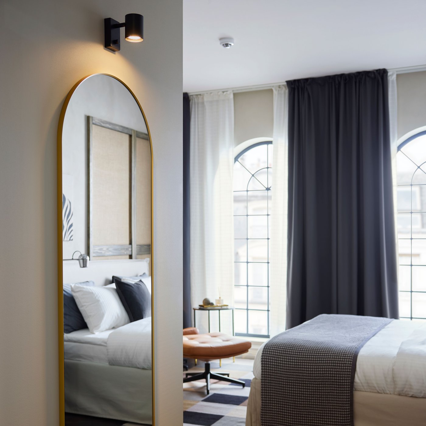 Ein grosser LINDBYN Spiegel mit goldfarbenem Rahmen, der oben abgerundet ist, spiegelt teilweise das aufgeräumte Bett eines Hotelzimmers wider.