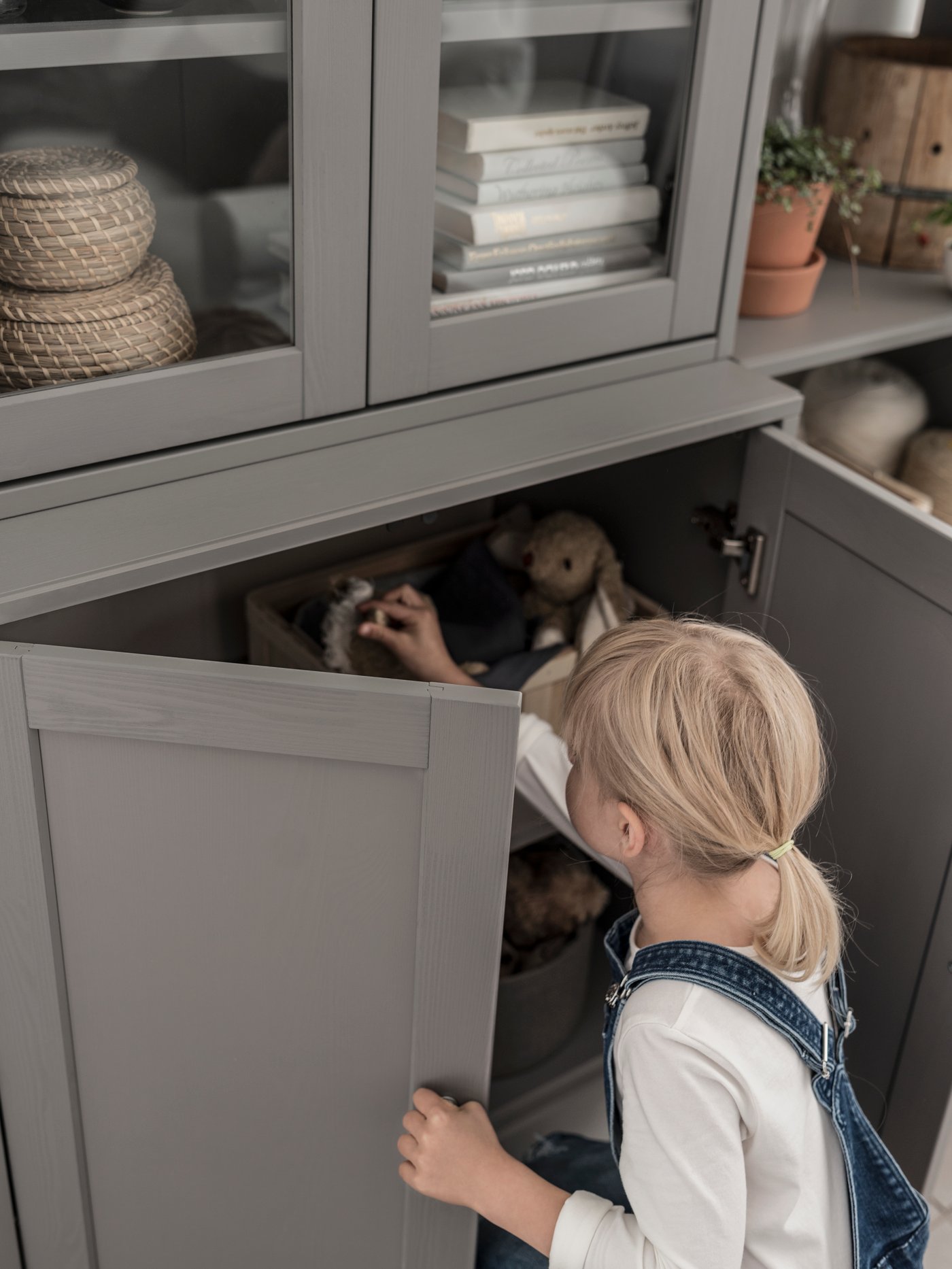 A child with denim overalls opens the bottom doors and reaches into a HAVSTA storage combination with glass doors.
