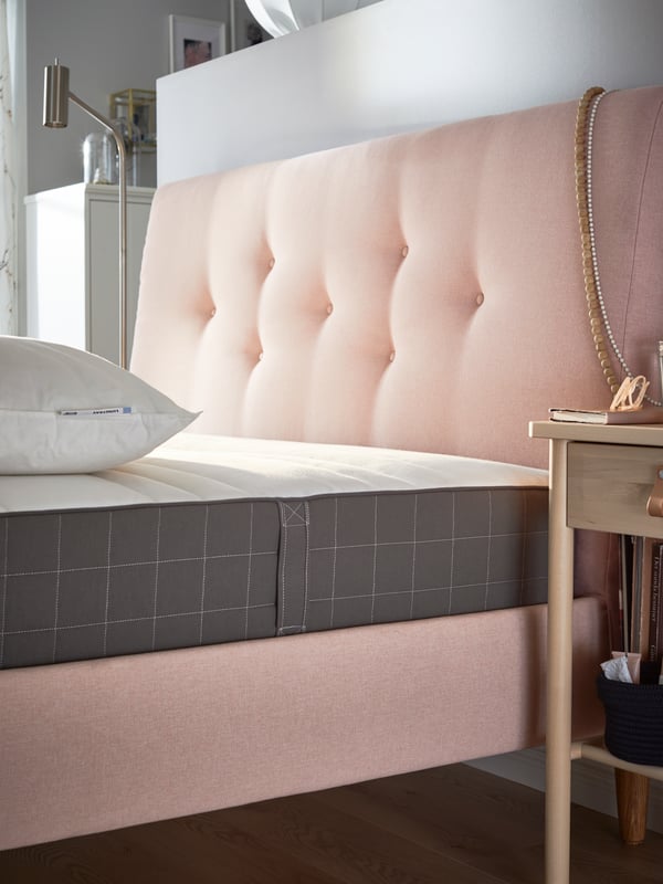 A mattress and pillow without covers on top of a pink upholstered bed with padded headboard.