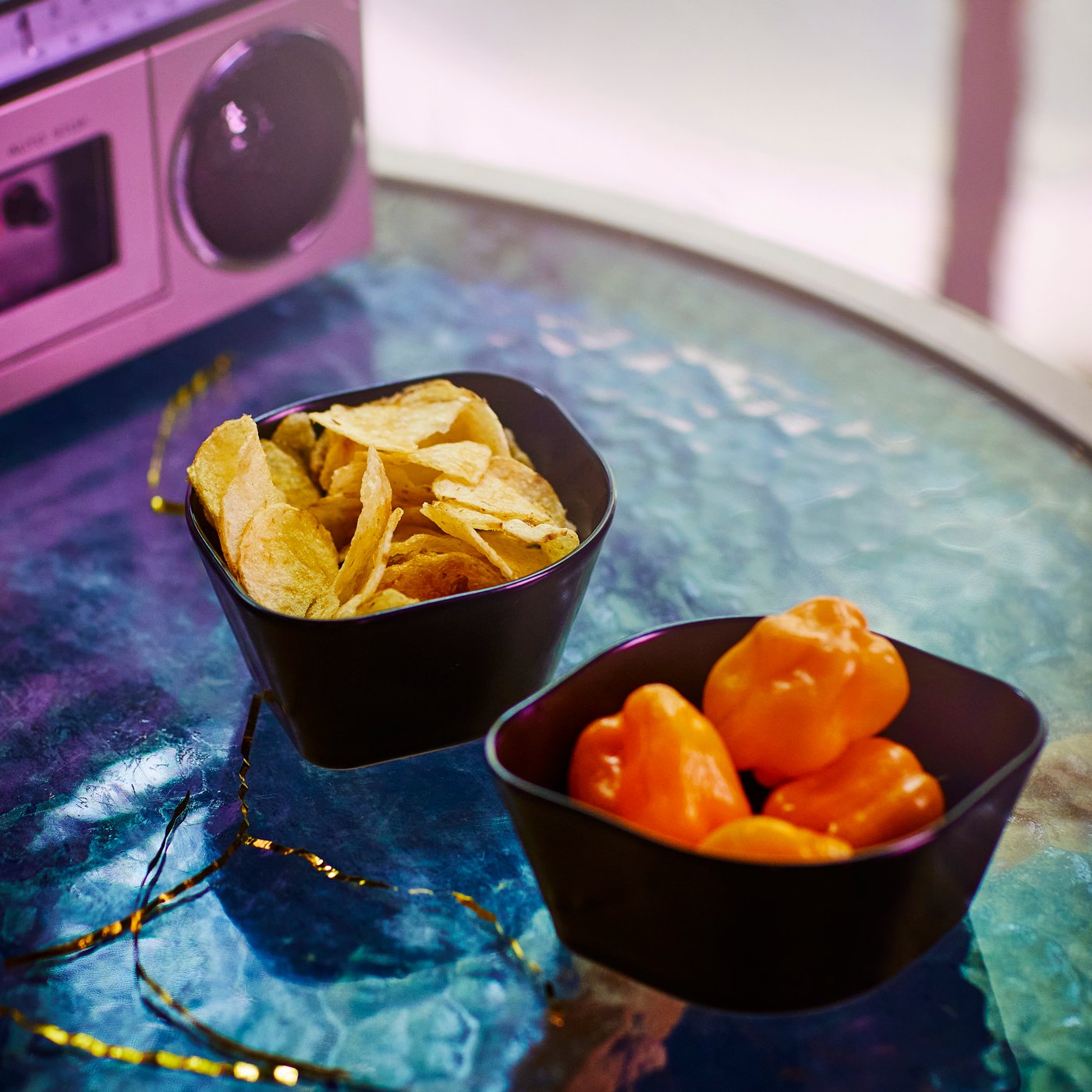 Two black ÖMSESIDIG serving bowls in stoneware filled with small peppers and nachos on a coloured glass table.