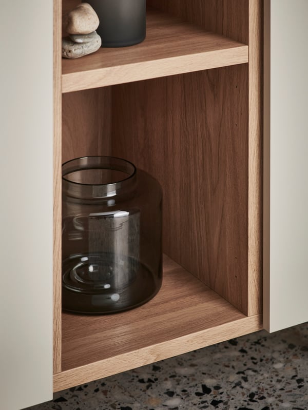 The open wood-effect section of a grey-beige LEDAMOT cabinet with a dark-grey glass vase and other decoration on its shelves.