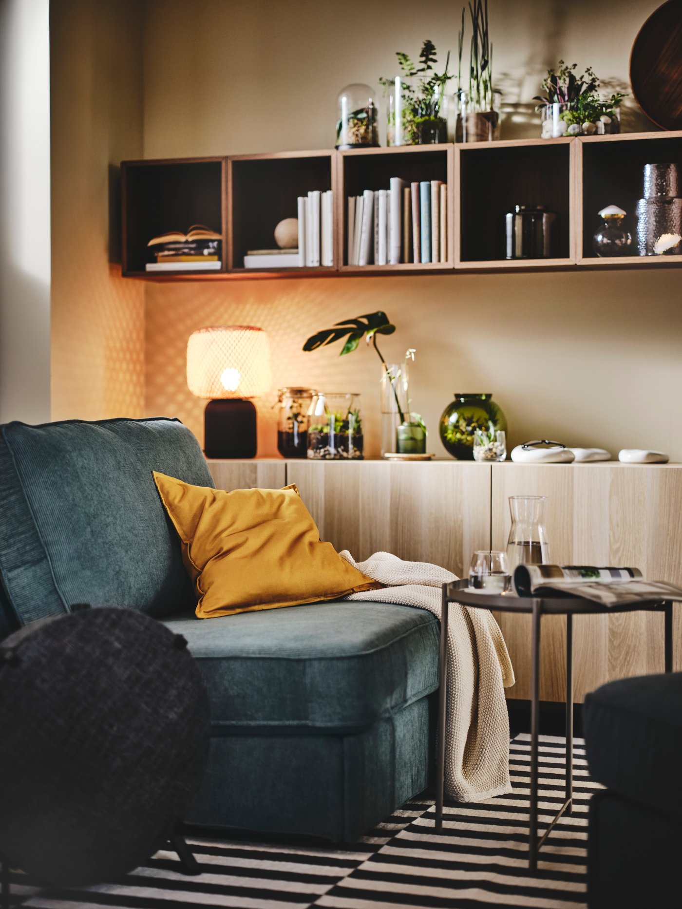 Un salon chaleureux avec un canapé vert foncé et un coussin jaune moutarde, un tapis rayé noir et blanc, une petite table avec un verre d’eau et des livres, et un meuble en bois rempli d’objets déco et éclairé par une lampe orangée.