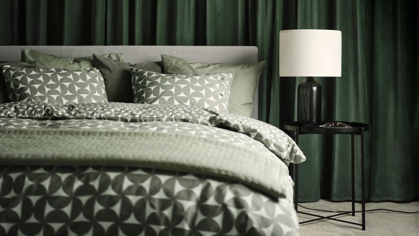 Una cama tapizada GLADSTAD con cuatro cajones, con ropa de cama  ÄNGSNEJLIKA color gris/verde.