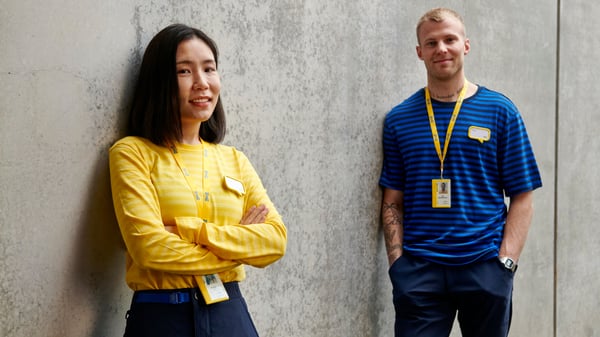 Dos colaboradores de IKEA están de pie frente a un muro de hormigón, la mujer lleva una camiseta amarilla y el hombre una azul.