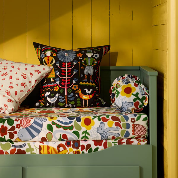 A multicolour TRATTKAKTUS cushion cover placed on a HEMNES day-bed frame with 3 drawers in grey-green against a yellow wall.