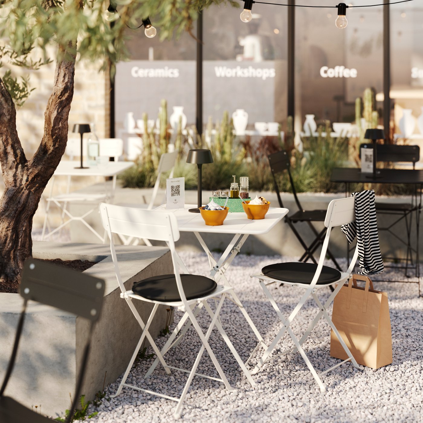 Im Außenbereich eines Cafés stehen auf einem Kiesbett neben einem Olivenbaum eine Reihe SUNDSÖ Klappstühle um einen Tisch herum.