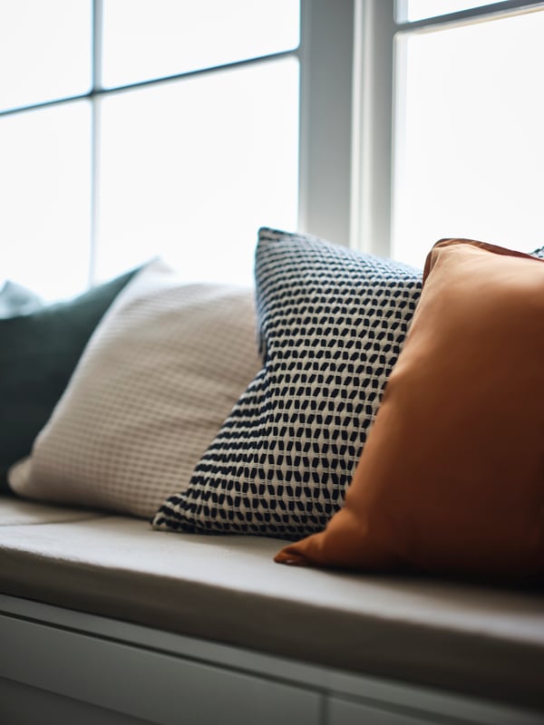 Four cushions, one with a KUSTFLY cushion cover, in assorted patterned and plain on a PLATSA storage bench against a window.