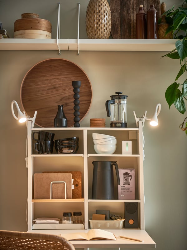 A white NORBERG wall-mounted drop-leaf table, its compartments filled with tableware and lit by two NÄVLINGE LED spotlights.