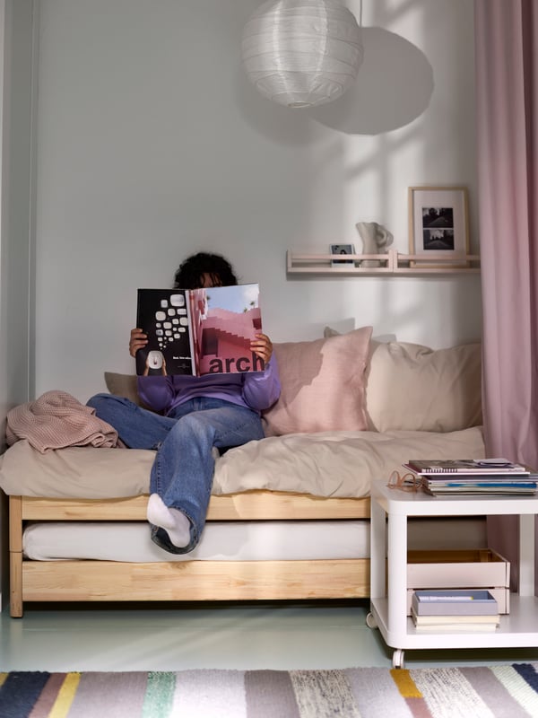 A woman sitting on two stacked UTÅKER pine beds reading a magazine, a white side table, a multicolor rug and a pendant.