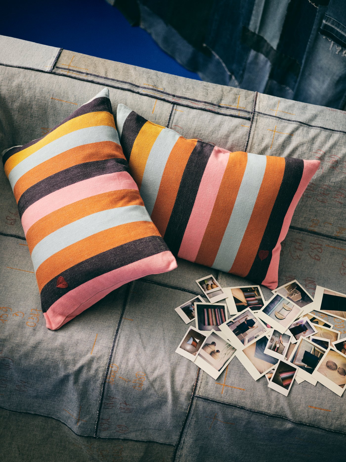 Dua kusyen dengan sarung kusyen MÄVINN berwarna-warni dengan perincian berbentuk hati bersulam tangan pada sofa dengan Polaroid.