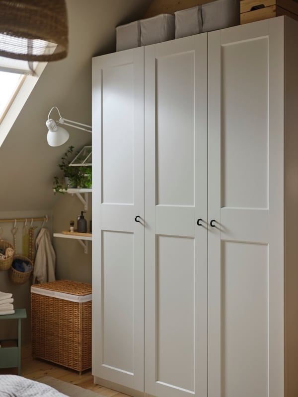 A white PAX/GRIMO wardrobe with three doors with a basket beside it in the corner of a room with a sloping ceiling.