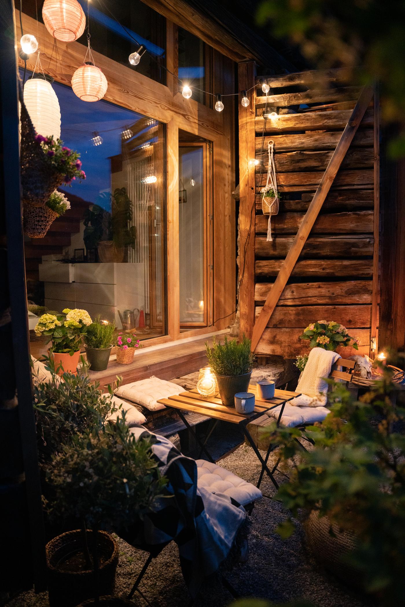 Stimmungsvolle Terrasse am Abend mit Lichterketten, Pflanzen und Holzmöbeln vor einem rustikalen Chalet.