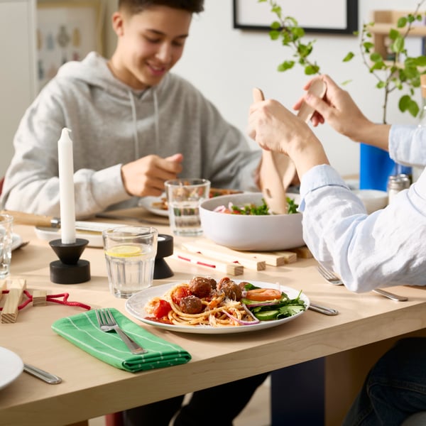Twee mensen zitten aan een eettafel, ze eten van witte GODMIDDAG borden en drinken water uit POKAL glazen.