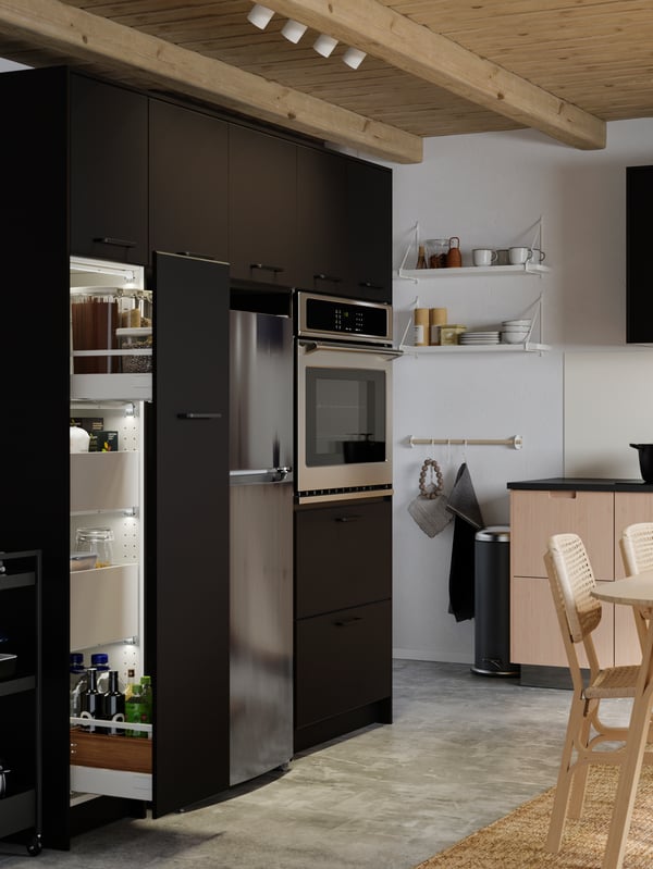 An anthracite pull-out pantry with bottles of oil and transparent containers with pasta, and two light bamboo chairs.