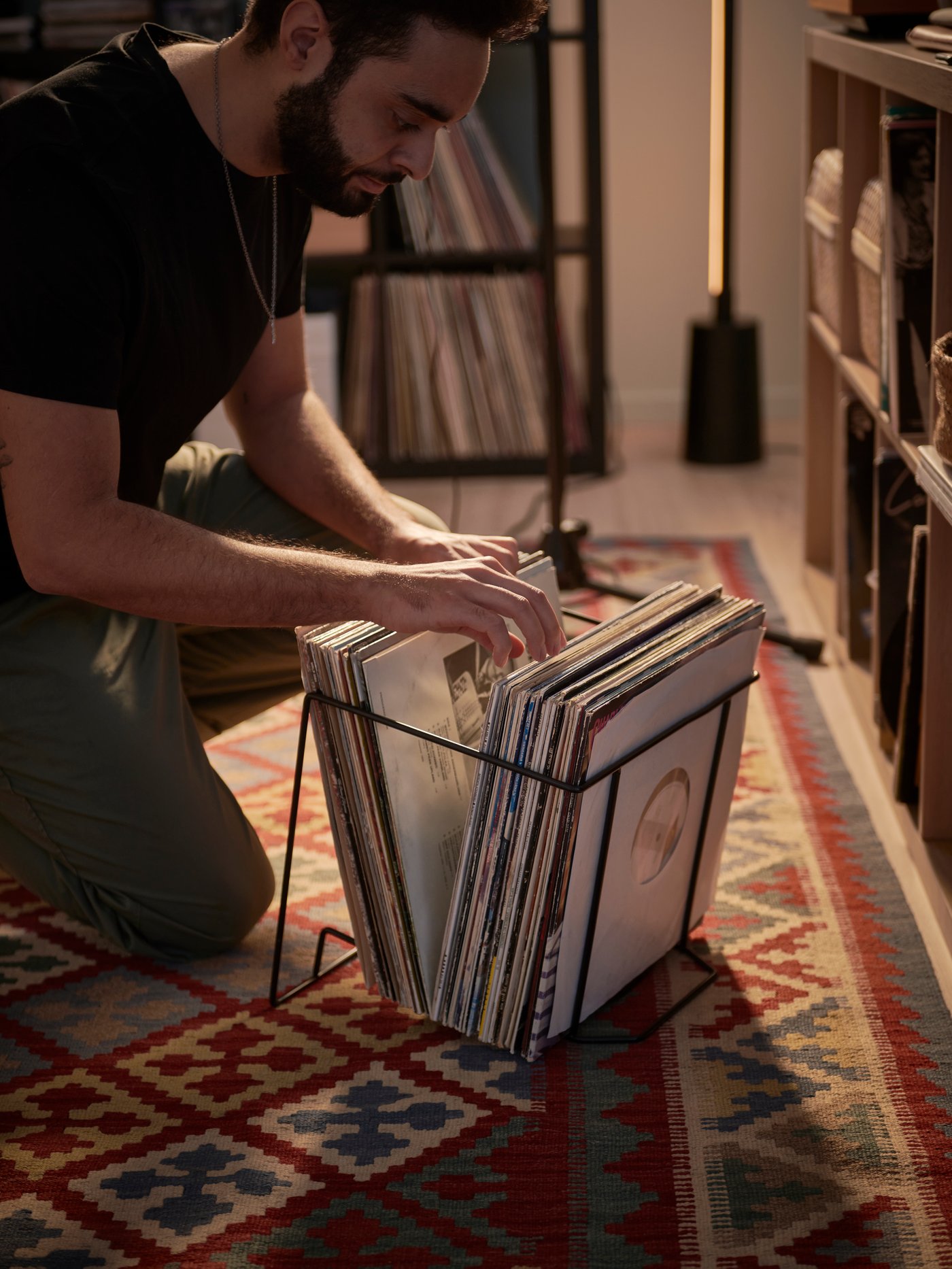 A man kneeling on a patterned rug flicking through vinyl records in the OBEGRÄNSAD record stand next to shelving units.