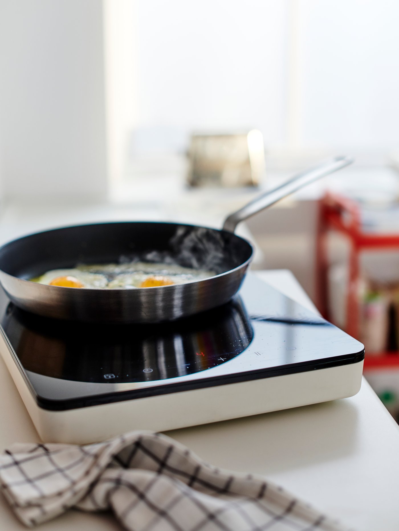 Permukaan atas kerja berwarna putih dengan hob aruhan mudah alih berwarna hitam/putih dan telur sedang digoreng di dalam kuali leper.