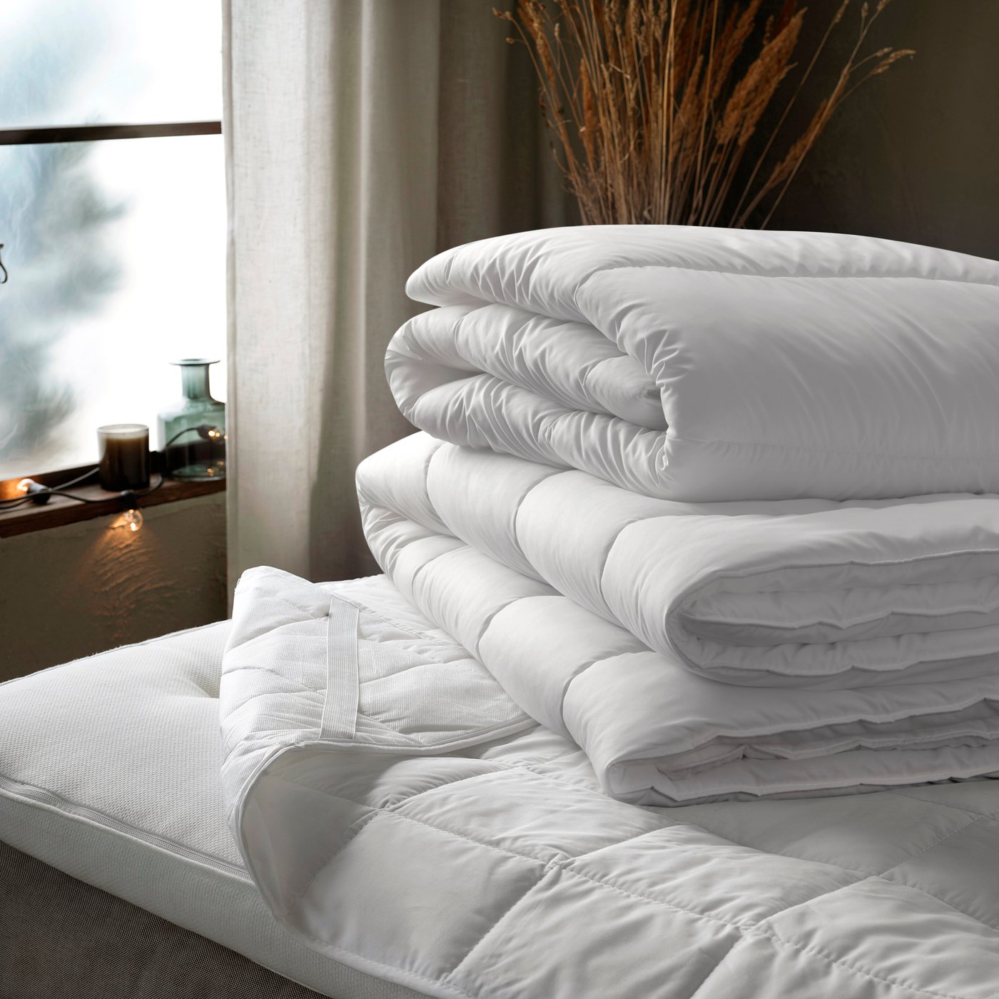 Two white, folded duvets in a pile on top of a bed with a mattress protector. A window with light curtains in background.