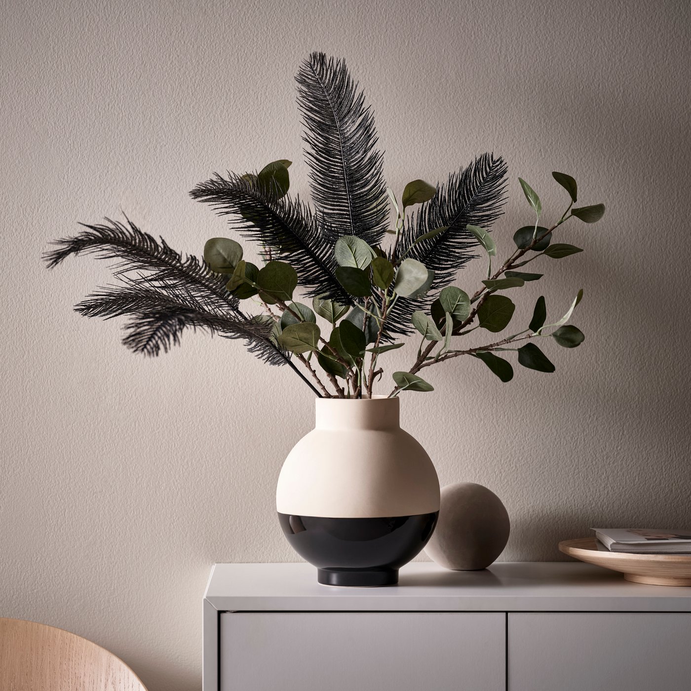 A black‑white GLANSGRAN vase filled with green SMYCKA artificial flowers and black SMYCKA plumes.