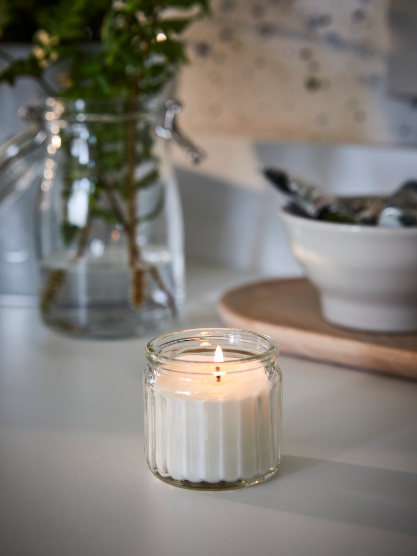 An ADLAD scented candle in a glass jar and a clear vase with leafy branches on a dining table.