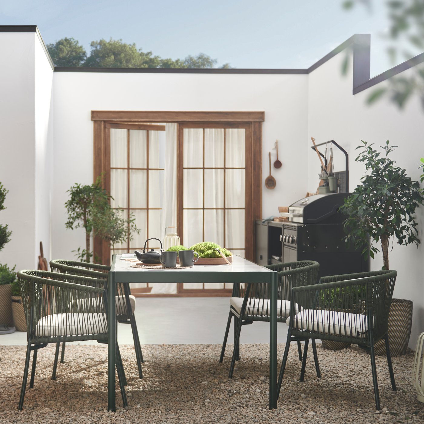 Leafy plants and IKEA SEGERÖN dark green/light grey table outdoor furniture on a bright, spacious, walled gravel-and-stone patio by a white, single-storey building.
