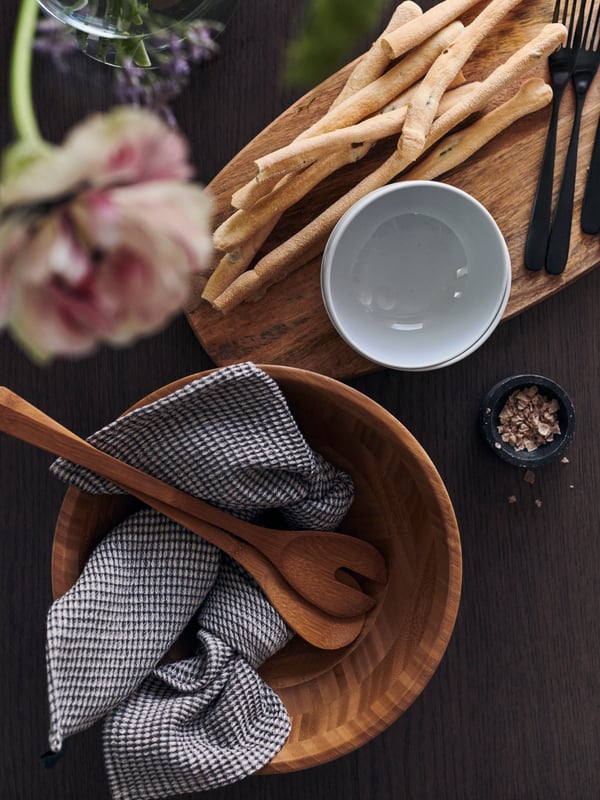 Un bol de service GRÖNSAKER vide et des ustensiles à salade avec une serviette de table à l’intérieur, à côté d’une planche à pain, des gressins et des fourchettes.