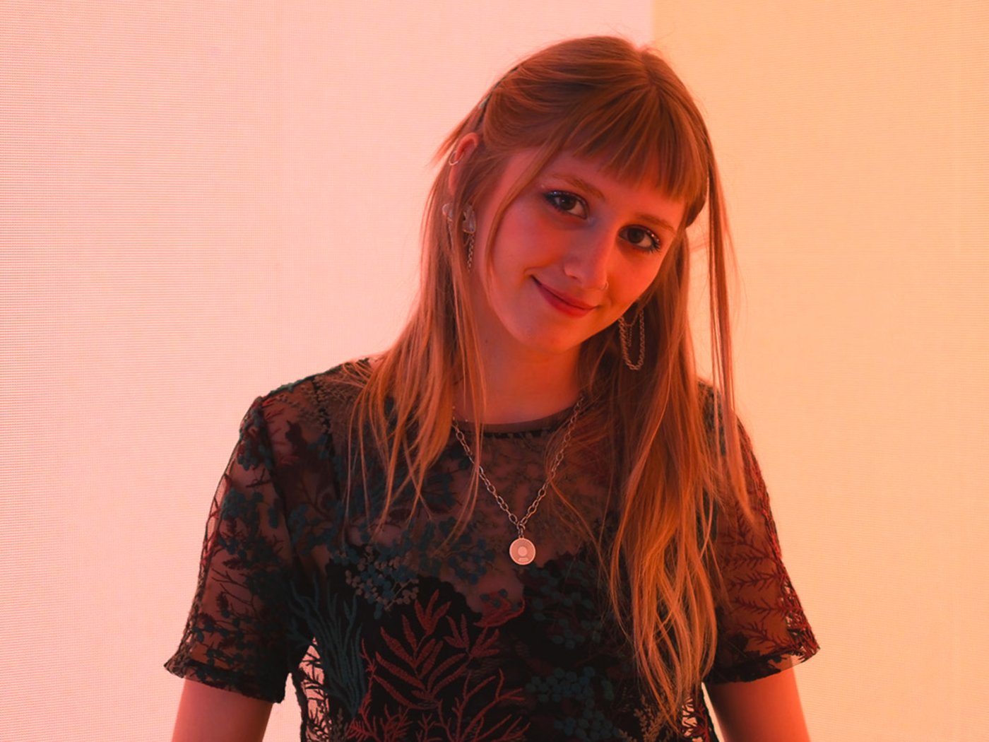 Rea standing on stage in a black t-shirt and necklace with a soft red colour as a background that comes from the digital display used on stage during Milan design week 2024 at the IKEA venue.