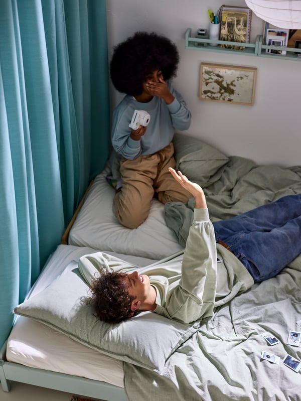 A man and woman on two beds with green BERGPALM and light gray/mélange SPJUTVIAL bed linen, by a turquoise curtain.