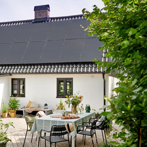 A sunny patio in front of a white house with dark gray roof tiles and solar panels on the roof. On the patio is a table with a light green tablecloth, set with glasses, a carafe, bread and flowers in a vase. Around the table are black chairs, and in the background is a bench with cushions and potted plants. Green bushes frame the picture.
