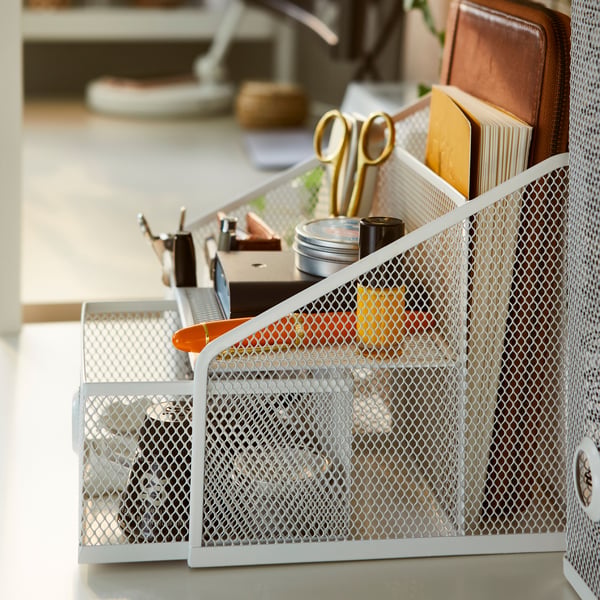 A DRÖNJÖNS desk organiser, holding many items such as a pen, a pair of scissors and a notebook, stands on a desk.