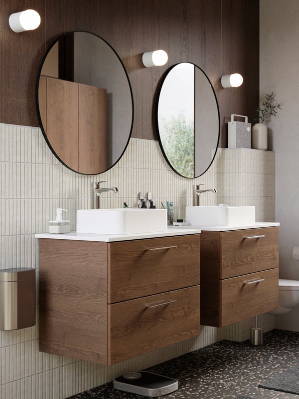 Two wooden washbasins, two large round mirrors, three wall lamps on the wall, beige tiles, dark terrazzo flooring.