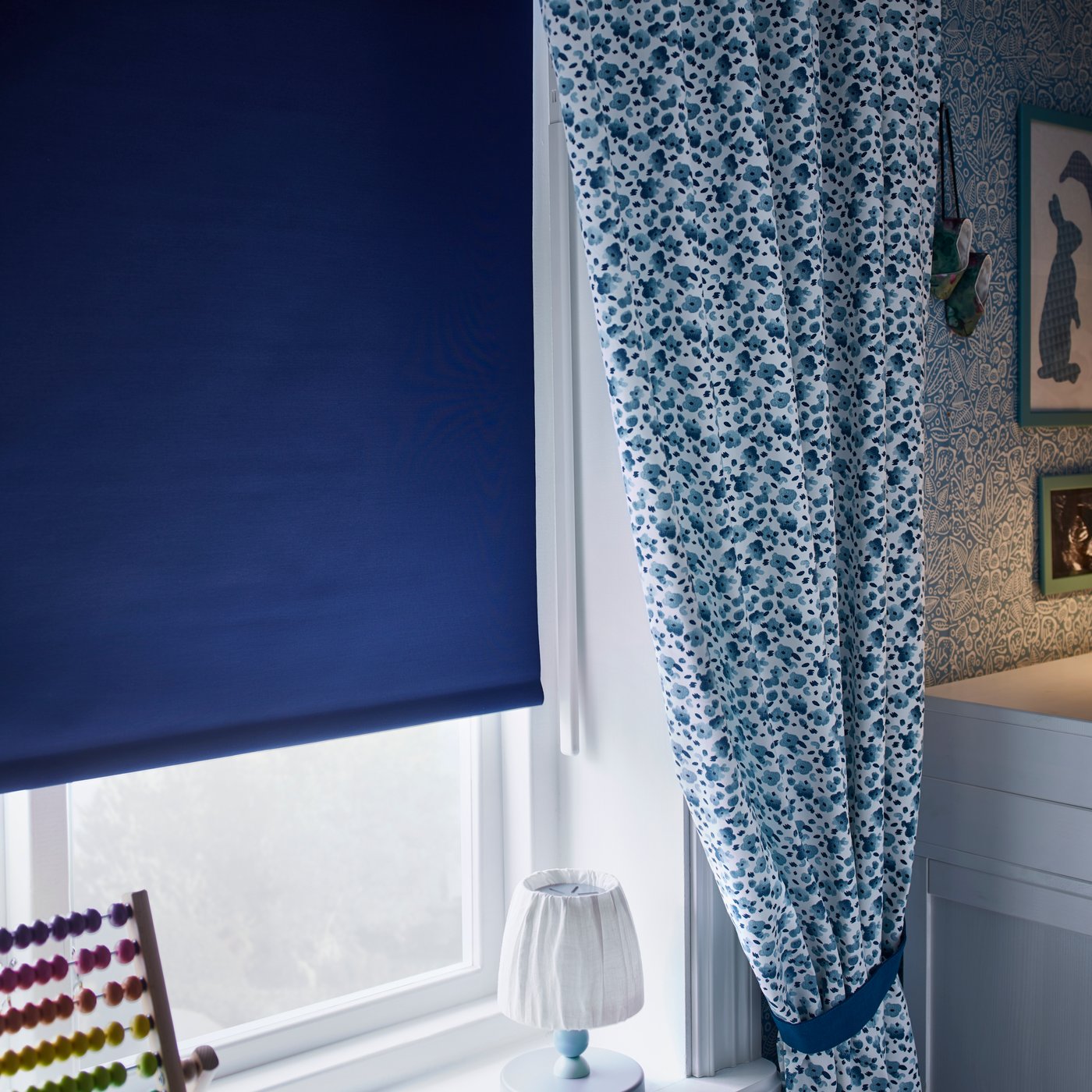 Vista de una habitaión infantil donde se aprecia una cortina roller FRIDANS  azul tapando una ventana de marco blanco. Al costado izquierdo de la ventada un juguete de madera IKEA y al otro costado  una cortina con diseño floreado de tonos azules.
