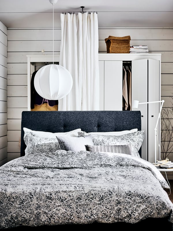 An IDANÄS upholstered bed covered in ÄNGSKLOCKA bed linen, with a bedside table, in front of a white DYTÅG curtain.