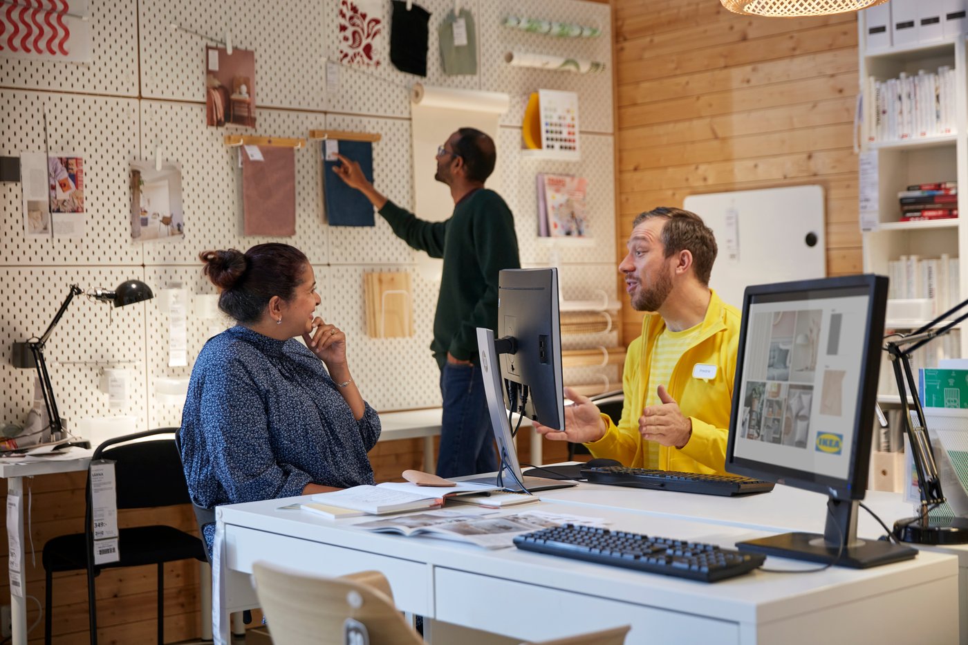 En kvinde sidder foran en IKEA medarbejder og taler mens en mand i baggrunden kigger på en væg hvor der hænger forskellige stilarter og materialeprøver.