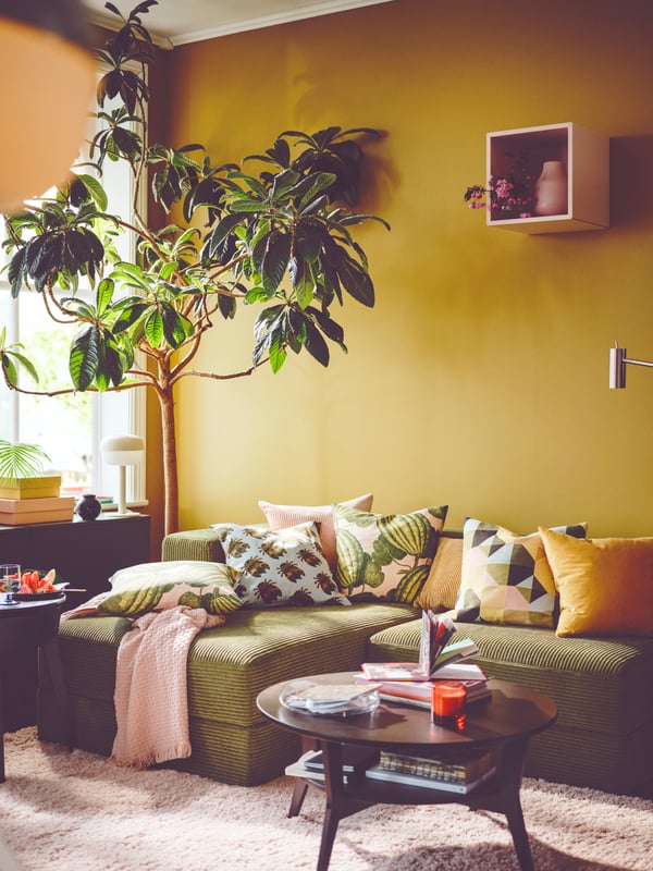 Eine senfgelbe Wand mit einem grünen JÄTEBO Sofa mit verschiedenfarbigen Sofakissen.